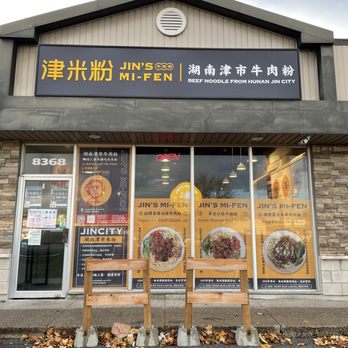 Jin’s Mi-Fen - The image shows the exterior of Jin's Mi-Fen, a restaurant located at 8368 in Markham, ON. The storefront has a large sign with Chinese characters, the restaurant name in English, and a description of beef noodle from Hunan Jin City. The glass windows display promotional posters and menu items, including images of beef dishes. Three wooden barriers are placed in front of the entrance, with fallen leaves on the ground. The building has a stone facade and a sloped roof. - Chinese Near Me - Markham, Ontario