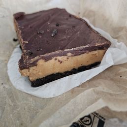 PB brownie - A layered dessert bar features a dark chocolate topping with a smooth surface and some small chocolate pieces, a caramel-colored middle layer, and a dark cookie crust at the bottom. It is placed on a piece of parchment paper on a textured surface, with a blurred background.