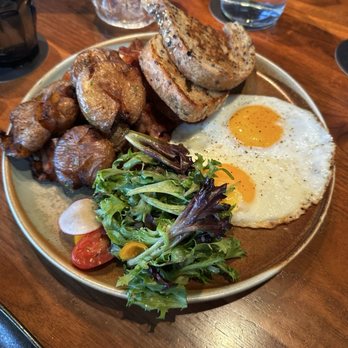 King Taps - A breakfast plate includes two sunny-side-up fried eggs, two slices of toasted bread, roasted potatoes, sausage pieces, and a mixed green salad with cherry tomatoes. The meal is served on a round ceramic plate placed on a wooden table, with a glass of water and a glass of dark beverage nearby. - Canadian (New) Near Me - Toronto, Ontario