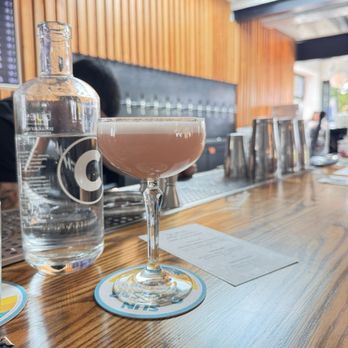 Chomp Kitchen and Drinks - A pink cocktail in a coupe glass is positioned on a round coaster on a wooden bar counter. Next to it is a clear water bottle with a black and white label. The background features wooden paneling, a black padded wall, and a row of upside-down glasses. Natural light enters from a skylight, illuminating the scene. - Burgers Near Me - Newport, Rhode Island