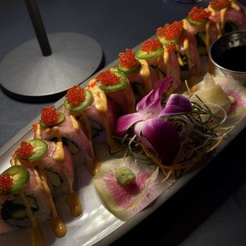 Ostras - Maki Tuna - A sushi roll features slices of cucumber, fish roe, and a drizzle of sauce on top, arranged on a white plate. Accompanying the roll are edible flowers, wasabi, and pickled ginger, with a dark dipping sauce in a small container. The setting appears to be a dimly lit restaurant table. - Asian Fusion Near Me - Des Plaines, Illinois
