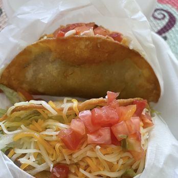 Papa Nachos - Pan Fried Crispy Taco Shells with shredded beef, cheese, lettuce & tomatoes - A taco with shredded cheese, lettuce, diced tomatoes, and a crispy taco shell, wrapped in paper. The taco appears to contain additional ingredients, possibly meat or beans, and is presented in a casual setting. The background shows a colorful tablecloth and a white container. - Mexican Near Me - Albuquerque, New Mexico