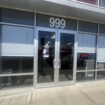 CubeSmart Self Storage - The image shows the exterior of CubeSmart Self Storage at address 999. The storefront has large glass windows and a glass door with a handle, with an open sign illuminated on the right side. The door has a QR code and a sign indicating contactless access. The building's upper part has windows with reflections of the sky and nearby structures. The sidewalk in front is visible, with a shadow cast on the ground. - Self Storage Near Me - Deer Park, New York