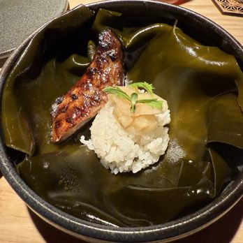 Ikigai - Kama toro - A black bowl holds a serving of white rice topped with pickled ginger and a small green herb. A grilled fish fillet with a glazed surface rests on large, dark green seaweed sheets that line the bowl. The presentation is simple and emphasizes traditional Japanese ingredients and preparation. - Japanese Near Me - Brooklyn, New York