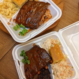 Two styrofoam containers contain barbecue dishes on a wooden table. The top container has a large piece of barbecue ribs with sauce, macaroni and cheese, a piece of cornbread, and a small portion of fried food. The bottom container has a barbecue rib, a piece of cornbread, coleslaw, and a smaller piece of meat. The presentation suggests takeout food from a barbecue restaurant or food truck.