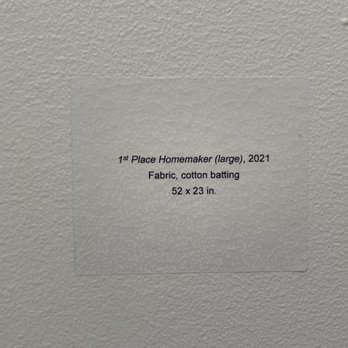 MOWA - St Kate - The image shows a wall-mounted label with printed text describing an art piece titled 'Place Homemaker (large)', created in 2021. The label states that the artwork is made of fabric with cotton batting and measures 52 by 23 inches. The label is affixed to a textured, light-colored wall. - Art Galleries Near Me - Milwaukee, Wisconsin