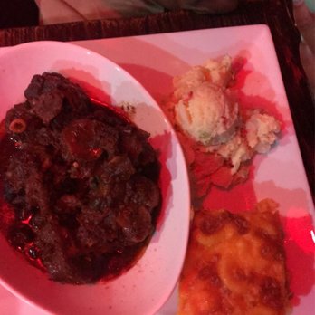 Sugarcane - Sugarcane 's Braised Oxtails with potato salad and Macaroni Pie - A square white plate contains three Caribbean-style dishes: braised oxtails in a dark sauce, a scoop of mashed potatoes with a small serving of ice cream on top, and a slice of baked lasagna with melted cheese. The plate is on a dark wooden table, and part of a person's arm and hand are visible in the background, suggesting a dining scene in a restaurant. - Caribbean Near Me - Brooklyn, New York