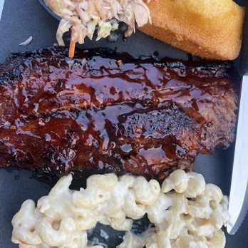 The Rusty Pig - A meal featuring a barbecue pork rib with barbecue sauce, macaroni and cheese, coleslaw, and a fried item, served on a black tray. - Barbeque Near Me - Geneva, New York