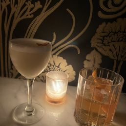 Two beverages (an old fashioned and the petro yuan) on a counter. A candle burns softly. Two beverages (an old fashioned and the petro yuan) on a counter. A candle burns softly.