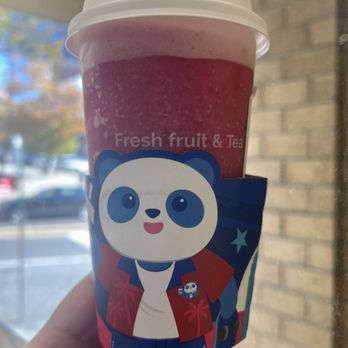 Tea By Do - Grape jasmine tea smoothie - A person holds a pink fruit and tea smoothie in a cup with a panda cartoon sleeve, outside near a window with a brick wall and trees in the background. - Bubble Tea Near Me - Flushing, New York