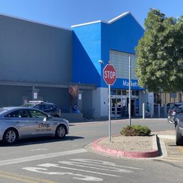 A Walmart Supercenter storefront with a parking lot, cars, and a stop sign in front. The building has a blue and gray facade with a few shoppers nearby.