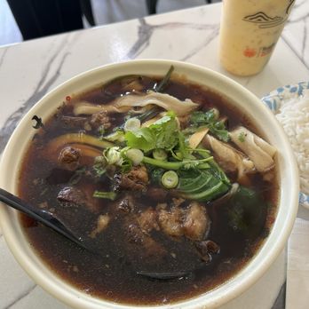 Yo Cafe - Braised Beef Noodles - A bowl of beef noodle soup with herbs, green onions, and vegetables, accompanied by a side of rice and a drink in a restaurant setting. - Chinese Near Me - Timonium, Maryland
