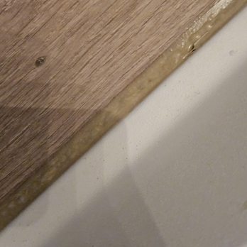 Anatom Restoration - Awful Grout job.  Doesn't match the floor or the tub.  And it's bubbled. - A photo showing the edge where a wooden surface meets a white wall, with bubbling and uneven sealant visible. - Damage Restoration Near Me - Centennial, Colorado