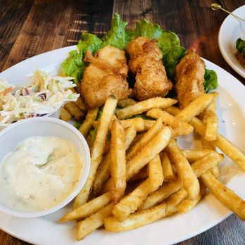 Rooster Run Cafe - Fish & Chips - A white plate holds fried fish fillets with a side of French fries, coleslaw, and a small container of tartar sauce. Lettuce leaves are placed under the fish. The plate is on a dark wooden table, with a partially visible sandwich and a bottle in the background. - Steakhouses Near Me - Hurricane, Utah