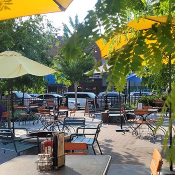 George's Local - Outside dining - The image shows an outdoor dining area with multiple tables and chairs, some with umbrellas providing shade. There are trees and greenery around the space, and string lights are visible hanging above. The setting appears to be part of a restaurant or cafe, with a paved surface and a fence enclosing the area. Vehicles are parked in the background, and a sign is visible among the trees. - New American Near Me - Cedar Falls, Iowa