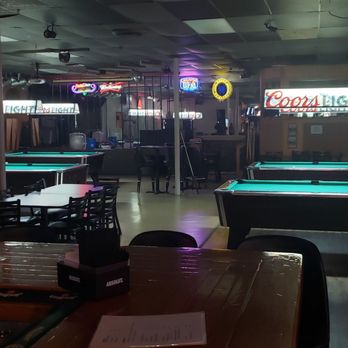 Maple Avenue Pub - Maple Ave Pub looks just as divey as you'd expect on the inside. Pool tables, pinball, and quasi-sketchy vibes - The image shows a bar with multiple pool tables illuminated by overhead lighting, neon signs including Coors Light, and various seating options such as chairs and tables. The space has a relaxed, dimly lit ambiance typical of a pub or billiards hall, with a bar counter visible in the background and decorative lighting fixtures hanging from the ceiling. - Pubs Near Me - Lisle, Illinois