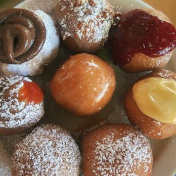 Jelly - One of each flavor - Donut Holes - A round plate holds multiple donut holes with various toppings and fillings. Some are dusted with powdered sugar, others have fruit preserves or chocolate icing, and one has a swirl of chocolate. The donut holes are arranged closely together, showcasing a variety of flavors and textures, on a neutral-colored surface. - New American Near Me - Denver, Colorado