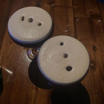Basso - Espresso martini for a night cap - Two coffee drinks with foam topping and coffee bean garnishes are positioned on a wooden table. The drinks are served in glasses with stems, and the foam appears smooth with a few coffee beans on top. The table surface is wooden with visible grain and some minor imperfections. The lighting is low, creating shadows and a cozy atmosphere. - Bars Near Me - Saint Louis, Missouri