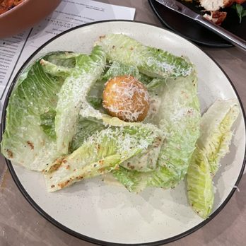 Lynette - ceasar salad - A Caesar salad with large romaine lettuce leaves, grated cheese sprinkled over the top, and a single crouton in the center. The salad is served on a white plate with a black rim, placed on a wooden table. In the background, there are other dishes and a menu visible, indicating a restaurant setting. - Beer Bar Near Me - Minneapolis, Minnesota