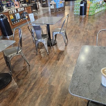 Raley's - Very accommodating seating area - The image shows an indoor grocery store with a seating area featuring metal chairs and tables. The tables have a gray, speckled surface, and the chairs are metallic with a simple design. The store has wooden flooring and shelves stocked with various products, including beverages and packaged goods, visible in the background. The area appears clean and organized, with some customers visible in the distance near the checkout counters. - Grocery Near Me - Placerville, California