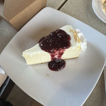 Toscano - Cheesecake with mixed berry compote - A slice of cheesecake topped with mixed berry compote is presented on a white square plate. The cheesecake has a smooth, creamy texture, and the berry topping is glossy and dark red. The plate is on a beige table, with a cardboard takeout box and other dishes visible nearby. The setting appears to be casual dining or takeout at a restaurant. - Italian Near Me - Draper, Utah