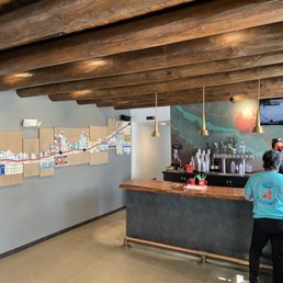 Cool vibe :) - The image shows the interior of a bar or cafe with a wooden ceiling made of large beams. A bar counter with staff and equipment is visible on the right side, with a person in a light blue shirt standing nearby. The wall behind the bar has a mural and a display of bottles and cups. On the left wall, there is a decorative art piece made of license plates and a cityscape. The space has a modern yet rustic design with warm lighting.
