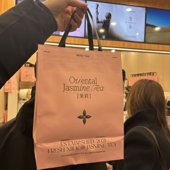 Molly Tea - A pink paper shopping bag from Molly Tea, labeled with Oriental Jasmine Tea and established in 2021, is held up by a person wearing a dark jacket. The bag features black handles and printed text, including product and brand information. The background shows a store interior with people, digital screens displaying images or videos, and a wooden ceiling. The scene suggests a retail environment focused on tea products. - Bubble Tea Near Me - Markham, Ontario