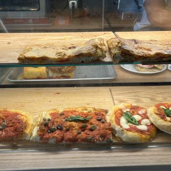 Pistacchio Pane e Caffe - Freshly made pizzas - The image features a glass display case with a wooden base, showcasing multiple slices of freshly baked pizza, including margherita and other varieties with toppings like olives and basil. The case is part of a food establishment, likely Pistacchio pane e caffe in Miami, FL, as indicated by the caption. Behind the glass, a person is partially visible, and a plate with a sandwich or similar item can be seen in the background. - Bakeries Near Me - Miami, Florida
