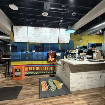 Laguna Burger - Counter area of Laguna Burger featuring digital menu boards displaying food options, a staff member working behind the counter, and a colorful mural with cityscape and geometric patterns on the wall. A small digital display stand and a branded mat are also visible. - Burgers Near Me - Albuquerque, New Mexico