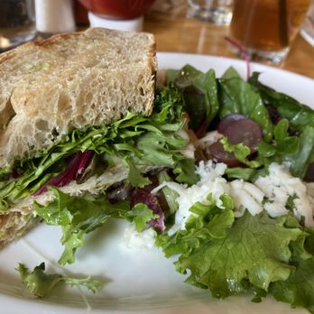 Bread+Salt - Chicken pesto sandwich - A sandwich with leafy greens and cheese on a toasted bread slice, accompanied by a mixed green salad with grapes and cheese, on a white plate. - Breakfast & Brunch Near Me - Frisco, Colorado