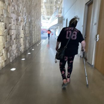 The Pit - Entry way from the south entrance - A woman with a cane and a shoulder bag walks through an indoor corridor featuring a stone wall on the left and wooden doors on the right. The corridor has a polished floor with embedded lights, and other people are visible further ahead, indicating a public or commercial space. - Stadiums & Arenas Near Me - Albuquerque, New Mexico