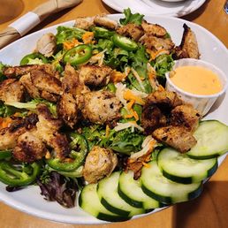 New Vegan Bahn Mi - A large mixed salad featuring grilled chicken pieces, sliced cucumbers, green peppers, shredded carrots, and leafy greens, served with a side of creamy dressing.