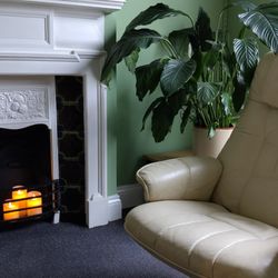 Creative Hypnosis & Hypnotherapy - A cream recliner chair sits beside a white-painted victorian fireplace with brown, green and blue tiles. Three candles glow warmly nearby.