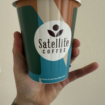 Satellite Coffee - Cafe con Leche - A person holds a Satellite Coffee paper cup with a colorful geometric design and a white plastic lid, against a plain wall background. - Coffee & Tea Near Me - Albuquerque, New Mexico
