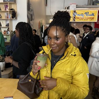 Drip Collective - Banana milk - A woman with braided hair in a yellow jacket holds a green drink with a straw at an indoor gathering with many people. - Coffee & Tea Near Me - Chicago, Illinois