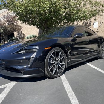 Premier Window Tinting - A black Porsche sports car with a sleek design and tinted windows is parked diagonally in a parking lot. The car's front is facing left, and it is positioned across multiple parking spaces. A tree with green foliage and a brick wall are visible behind the vehicle, with sunlight casting shadows on the ground and the car's surface. - Home Window Tinting Near Me - Reno, Nevada