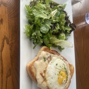 Perk Up Kitchen - Croque Madame with Mixed Greens - A rectangular white plate holds a breakfast dish with a fried egg on top of toast, garnished with herbs. Accompanying the dish is a side salad of mixed greens, including lettuce and spinach, arranged on the same plate. The setting appears to be a wooden table with glasses of water nearby. - Breakfast & Brunch Near Me - Bloomington, Minnesota