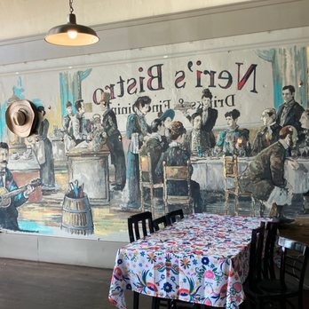 Neri's On the Square - Interesting wall mural. - The image features a large mural painted on a restaurant wall, illustrating a busy dining scene with multiple people seated at tables, some standing, and musicians performing. The mural includes detailed figures of men and women, with some engaged in conversation and others enjoying music. In front of the mural, there is a table covered with a colorful, floral-patterned tablecloth and surrounded by black chairs. Additional decor includes a hat hanging on a lamp post and a dress with floral embroidery hanging on the right side. The setting appears to be inside a casual dining restaurant with a vintage or rustic ambiance. - New American Near Me - Graham, Texas