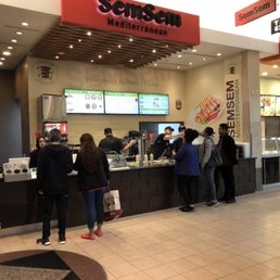 SemSem with the theater entrance to the left. - Inside a shopping mall, a SemSem Mediterranean restaurant counter features digital menu screens displaying food options. Several customers are lined up, ordering or waiting for their food. The counter area includes a beverage refrigerator and food display case. The restaurant's signage and branding are visible, with a modern interior design and a tiled floor.