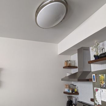 Inca Electric - This is the old light fixture that Inca Electric removed. - The image shows a modern kitchen with a white ceiling and walls. A round, flush-mounted ceiling light fixture with a metallic trim is visible. To the right, there are wooden shelves holding jars, a potted plant, and other small items. Below the shelves, a stainless steel range hood is installed above a stove, with a coffee maker and kettle on the countertop. The kitchen has a clean, minimalist design with functional appliances and decor. - Electricians Near Me