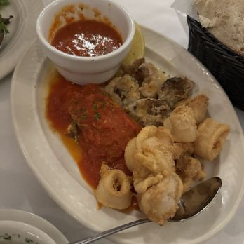 Gino's Restaurant - Hot Antipasto - A white oval plate holds fried calamari rings with a side of tomato sauce garnished with herbs. A small white bowl of soup or sauce is placed on the plate, and a lemon wedge is nearby. In the background, a basket contains slices of bread, and part of a glass is visible. The setting appears to be a restaurant table with a white tablecloth. - Pizza Near Me - Brooklyn, New York