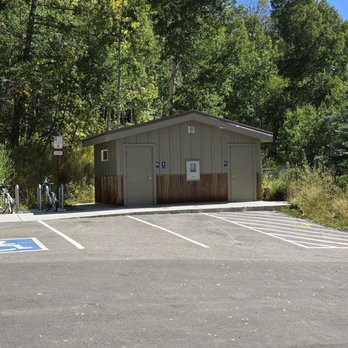 booth lake trail - Very clean restroom.  Modern convenience.  Diaper changing station - A small restroom building with a gray exterior and wooden accents is situated in a parking lot, surrounded by trees and under a bright blue sky. - Hiking Near Me - Vail, Colorado