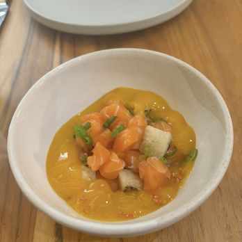 Tabú - A small, white bowl on a wooden table contains diced salmon, white fish, and chopped green onions in a yellow sauce or puree. The background shows an empty oval plate and a glass with a beverage. The setting appears to be a restaurant or dining area. - New Mexican Cuisine Near Me - Brooklyn, New York