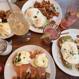 Lake Benedict - Three plates of eggs Benedict with potatoes, two pink drinks with lime wedges, a glass of white wine, and a small cup on a wooden table.