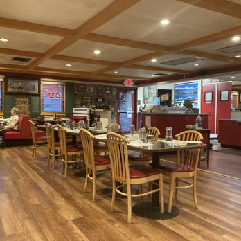 Namaste - Namaste.  Interior. - The image shows the interior of a restaurant with a wooden floor and a ceiling with a grid pattern. Several tables with chairs are arranged throughout the space, some with table settings. There are a few patrons seated, including a man and a woman on the left side. The walls are decorated with framed pictures and windows with blinds. The background includes a counter area with a digital screen and a kitchen section partially visible. The lighting is warm and evenly distributed. - Indian Near Me - Rio Rancho, New Mexico
