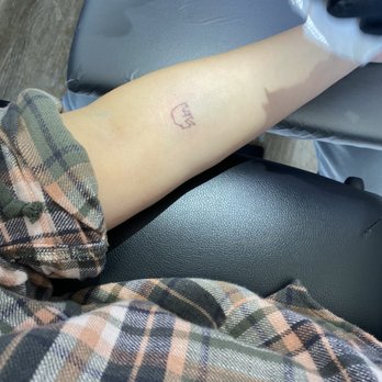 Kunce Tattoo - Tattoo - A person's forearm shows a small, simple tattoo of a bear outline, with the person wearing a plaid shirt and sitting on a chair. - Tattoo Near Me - Chicago, Illinois