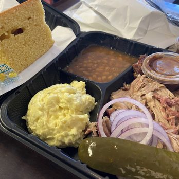 Black Bloc BBQ - Omg - A barbecue meal served in a black plastic tray includes pulled pork with sliced onions, a pickle, mashed potatoes, baked bread, baked beans, and barbecue sauce in small containers. The tray is placed on a table with paper and other items visible in the background. - Barbeque Near Me - Winthrop, Maine