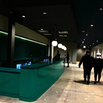 Ramova Theatre - upstairs bar - The image shows an indoor space with a green bar or reception counter, illuminated by hanging spherical lights. Several people are walking or standing in the background, with some engaged in conversation. The ceiling has multiple small recessed lights, and the floor appears to be concrete or similar material. The setting suggests a modern, possibly commercial venue such as a bar, lounge, or event space. - Cinema Near Me - Chicago, Illinois