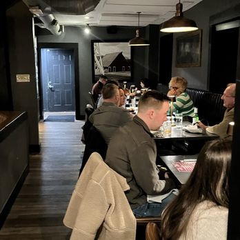Nyla's Restaurant - Inside seating - The image shows an indoor dining area of a restaurant with dark-colored walls and ceiling. Pendant lights hang from the ceiling, illuminating the tables where multiple patrons are seated. The patrons are engaged in eating and conversation, with some using their phones. The decor includes framed artwork on the walls and a black door at the back. The setting appears cozy and modern, with a mix of casual and semi-formal dining atmosphere. - Cocktail Bars Near Me - Westfield, Indiana