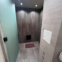 View from waiting area into roomy spray area - A shower stall features gray tiled walls and floor, with a frosted glass door on the left side. The ceiling has recessed lighting, and a small black vent is visible on the back wall. A red bath mat is placed on the shower floor. To the right, there is a gray textured wall with a sign and small drawers or compartments below it.